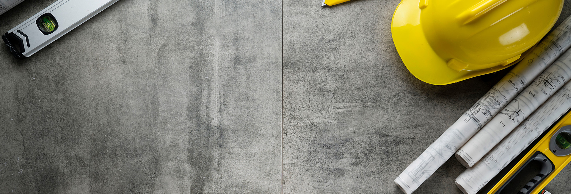 Baupläne, Wasserwaagen und gelber Schutzhelm auf grauem Betonboden – Symbolbild für Planung und Handwerk.