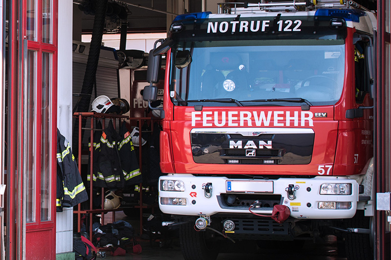 Feuerwehrfahrzeug in einer offenen Fahrzeughalle mit Einsatzbekleidung und Helmen an der Wand.