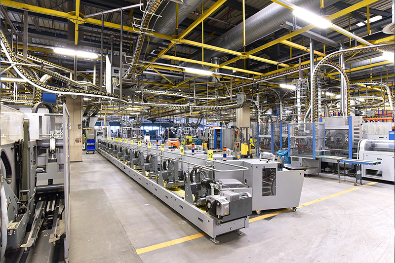 Moderne industrielle Fertigungshalle mit automatisierten Produktionsanlagen, Fördertechnik und zahlreichen Rohrleitungen unter der Decke.