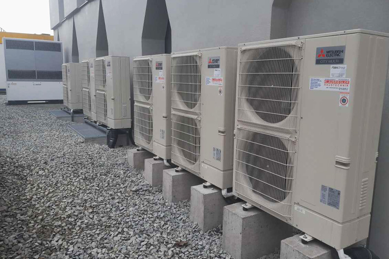Außenklimaanlagen der Marke Mitsubishi Electric, auf Betonfundamenten montiert, nebeneinander auf einem Schotterboden.