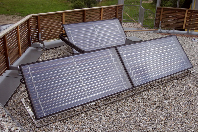 Solaranlage bestehend aus zwei Solarkollektoren, die auf einem Schotterbett auf einem Dach montiert sind, mit einem Holzgeländer im Hintergrund.